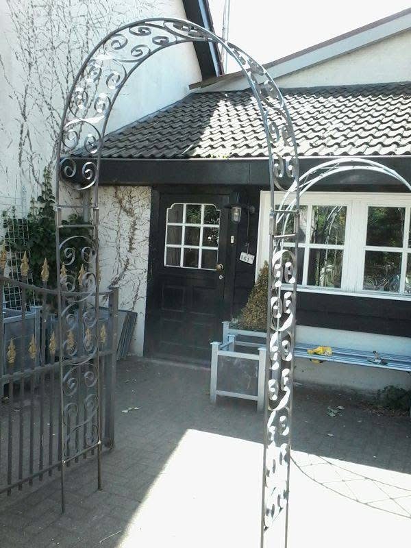 Handgesmede rozenboog met sierkrullen en gepunte spijlen, opgesteld bij entree